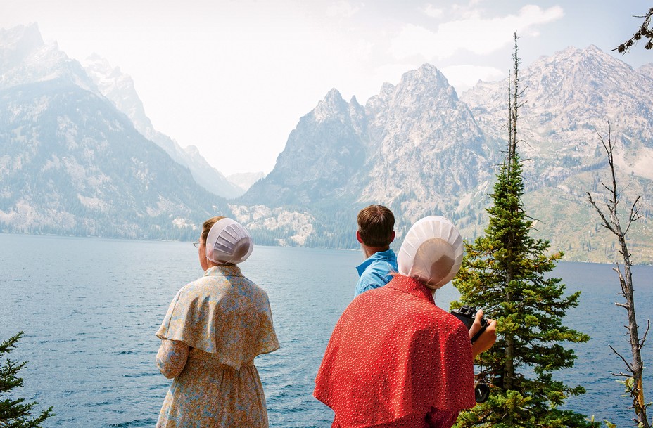photo of two women in Mennonite-style head coverings and dresses in front of lake with mountain behind