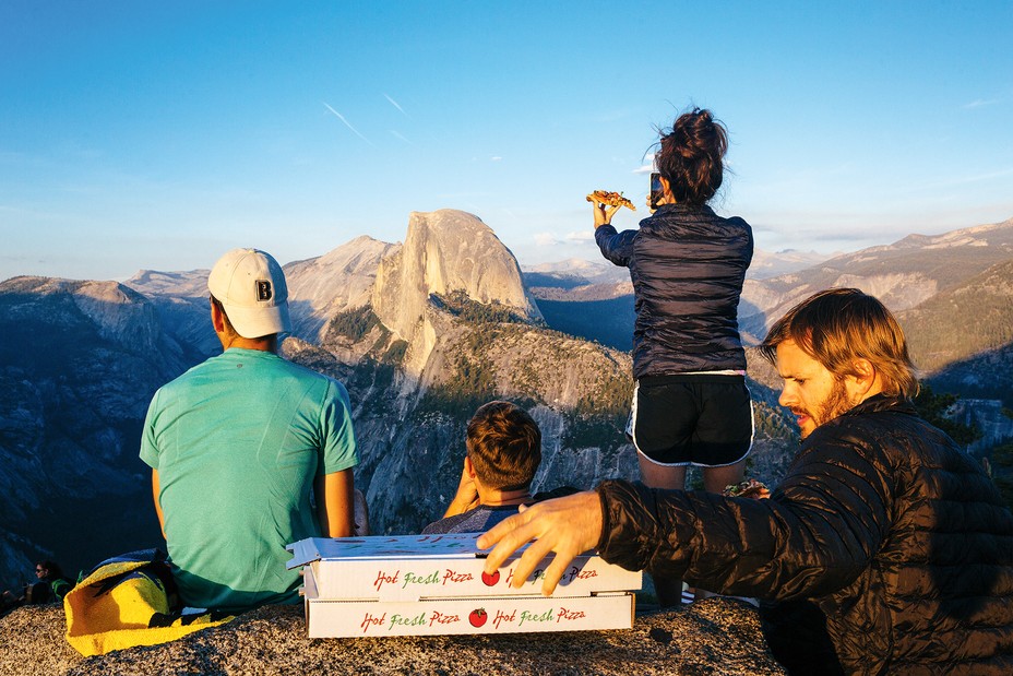 photo of people sitting with two pizza boxes, with woman standing taking photo of slice of pizza in front of view of Half Dome