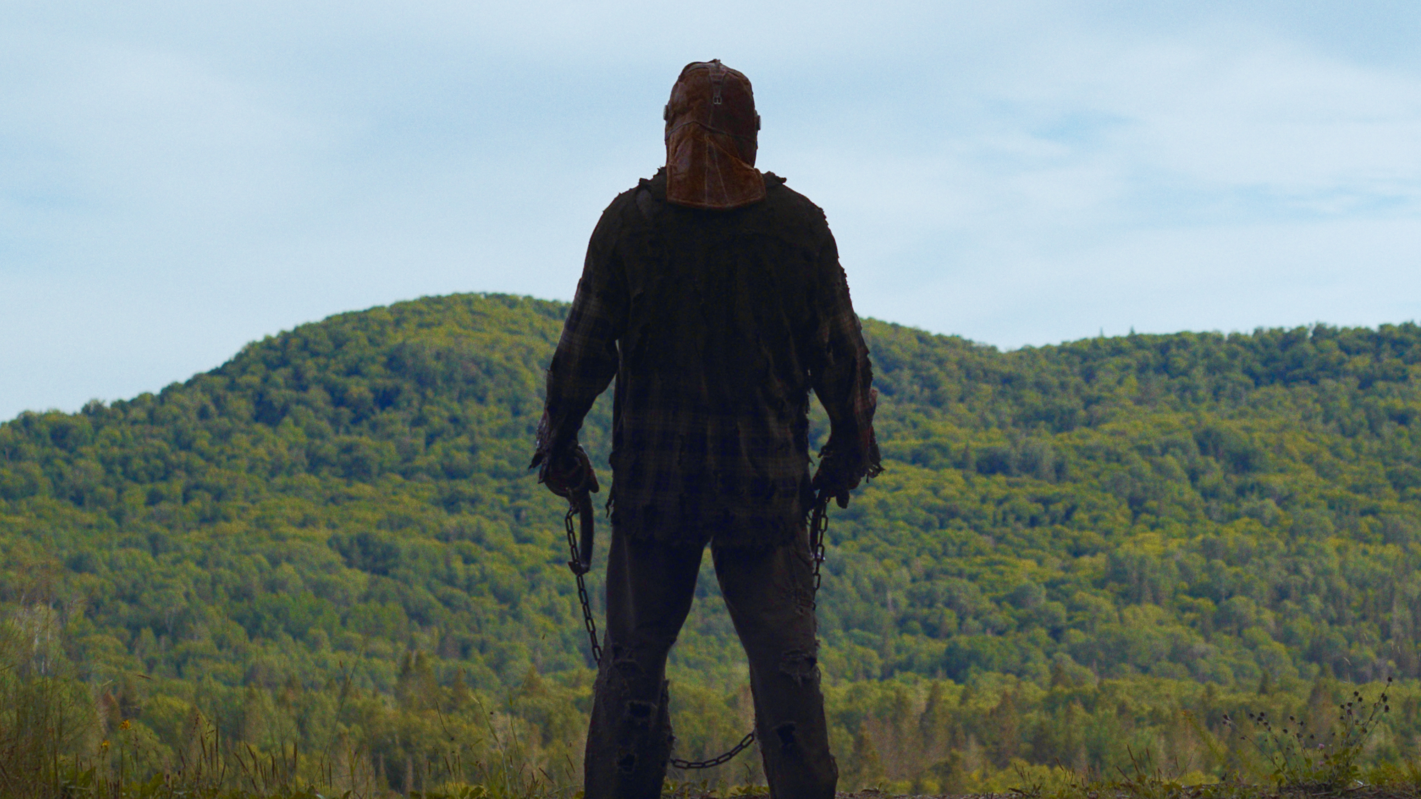 A masked figure holding a chain looks into the woods