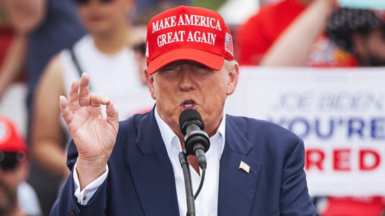 Wearing a red MAGA hat and a blue suit with a flag lapel pin, Trump speaks into a microphone at his recent rally in Las Vegas