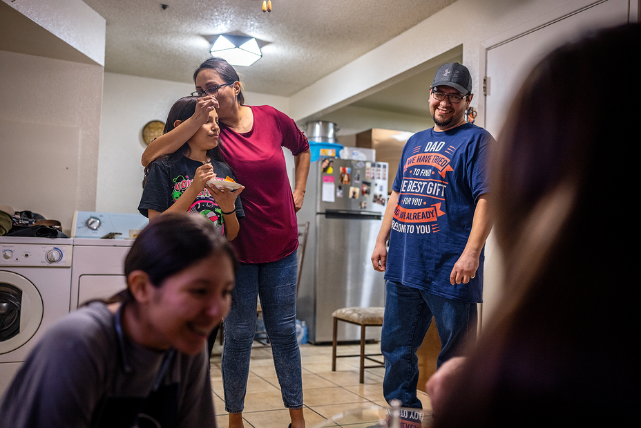 photo of standing woman kissing child on side of head with other family members smiling in tiled room with refrigerator, washer, and dryer