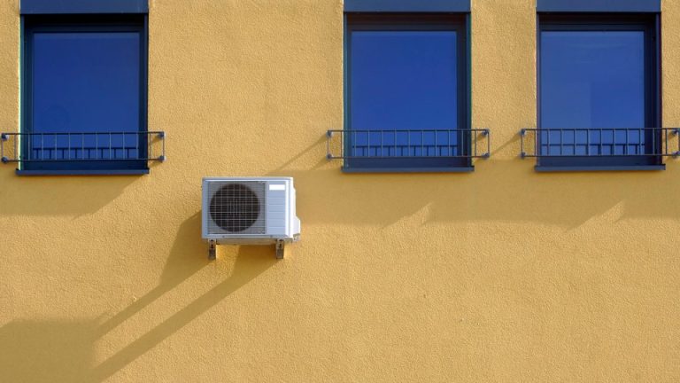 An AC unit mounted on a yellow wall that has three blue windows