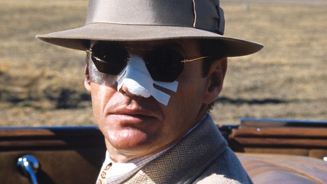 A movie still of Jack Nicholson, playing private eye J. J. Gittes in Chinatown (1974), in a convertible car with the top down, wearing a fedora and sunglasses with his nose covered in white medical tape.