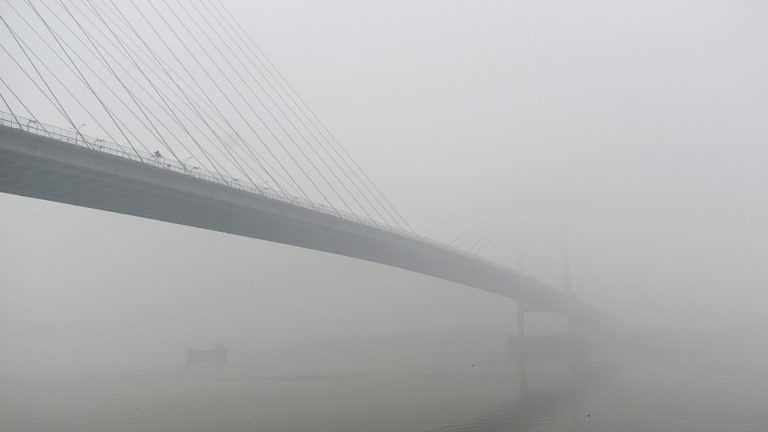 A bridge stretches over a wide river in India in thick fog.