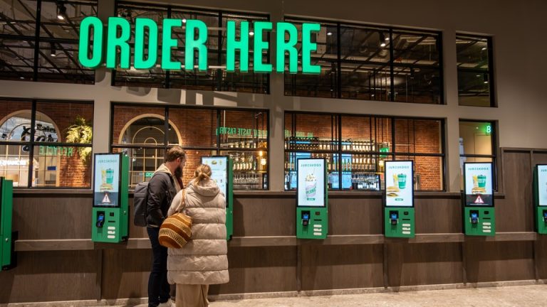 Two people stand in front of a self-checkout kiosk under a sign that says "ORDER HERE" at an airport