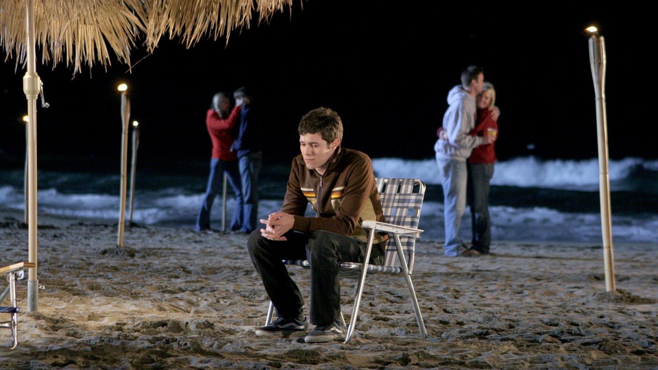 Seth Cohen sits alone on a beach chair while couples canoodle in the background