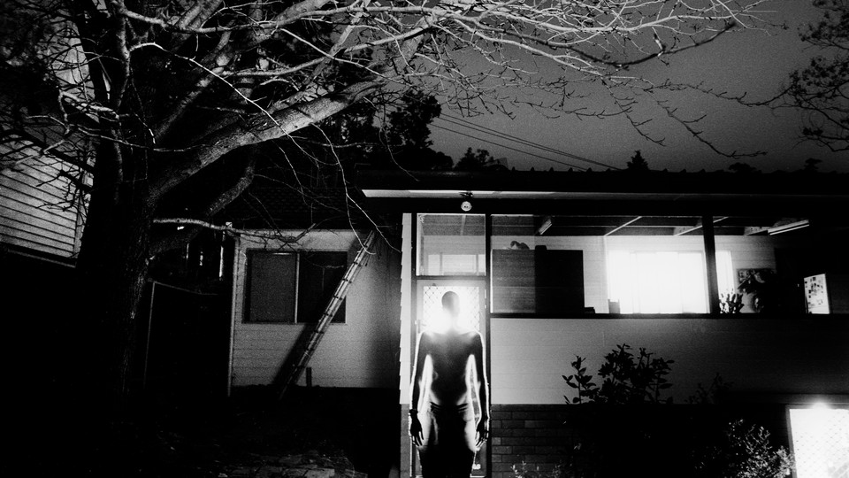 a man standing in darkness before a house, glowing from within