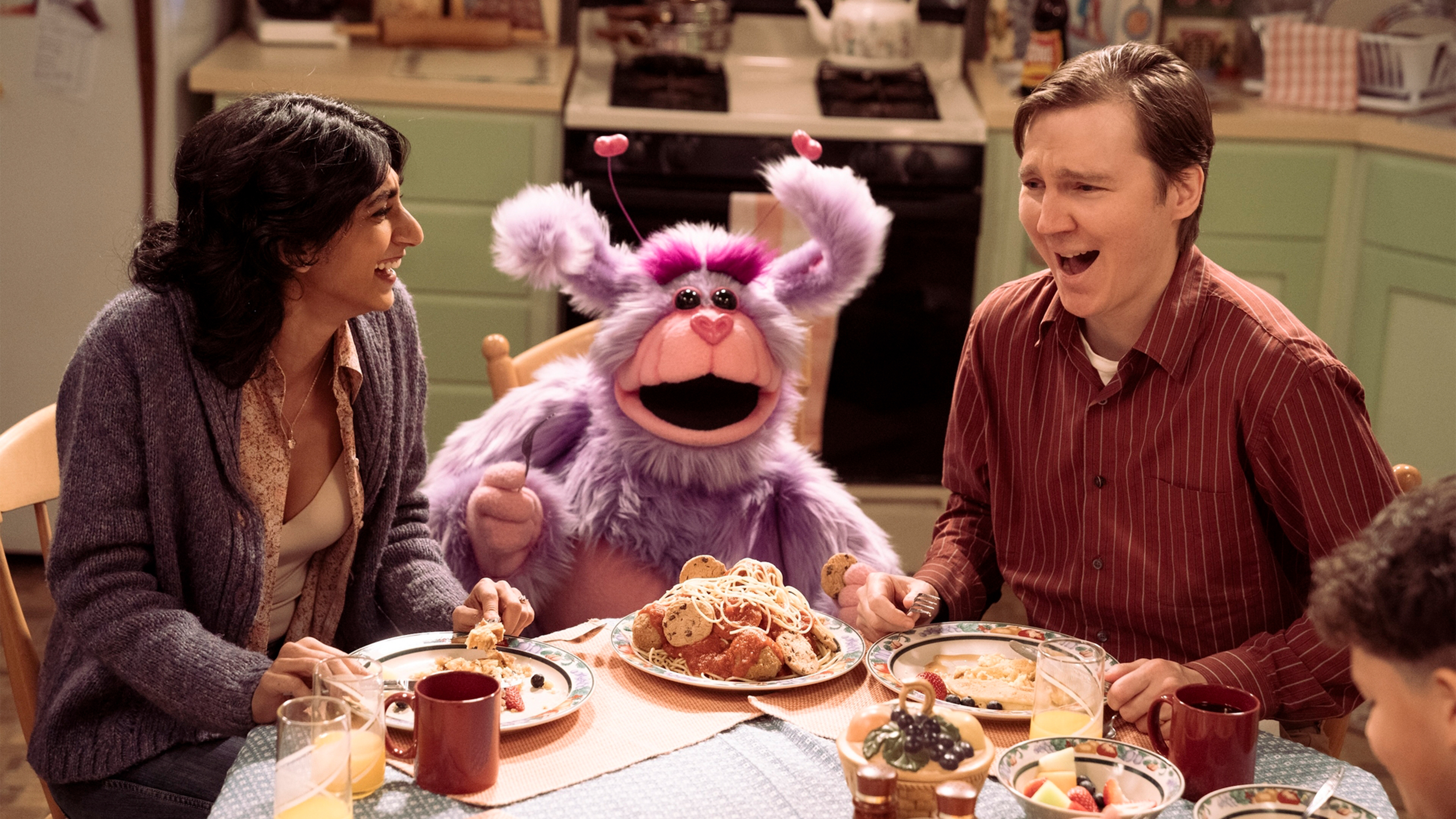 Two people laugh at a table and a puppet creature sits between them