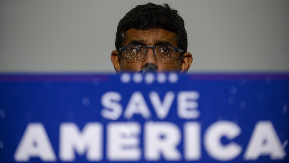 Dinesh D'Souza stands behind podium that reads: Save America. His glasses are spotted with rain.