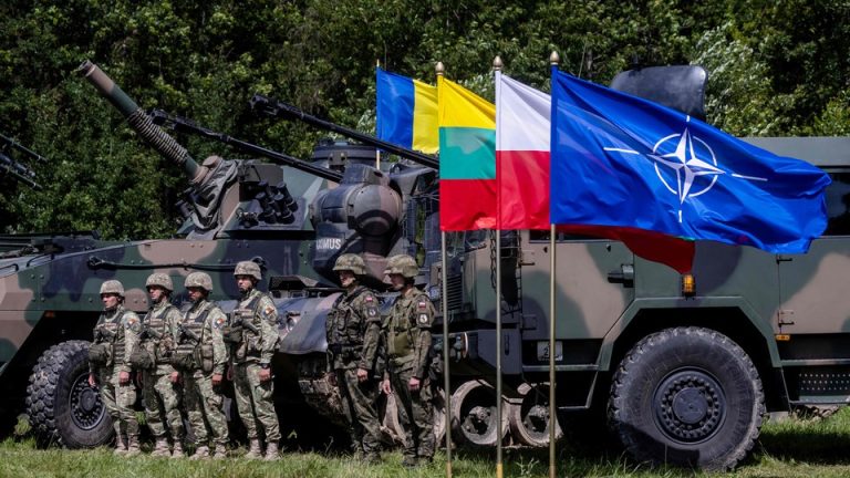 Soldiers stand next to tanks and the NATO flag