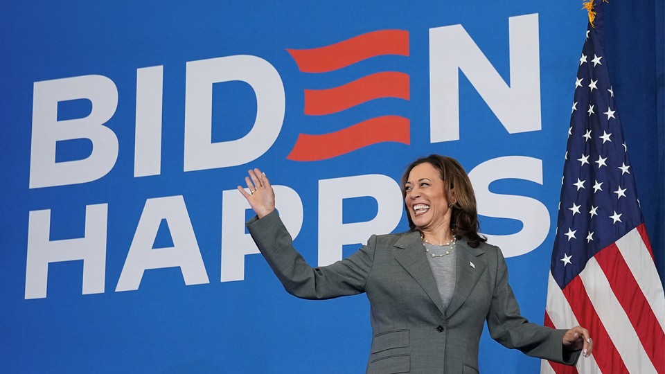 Kamala Harris waving happily in front of a BIDEN HARRIS sign and an American flag