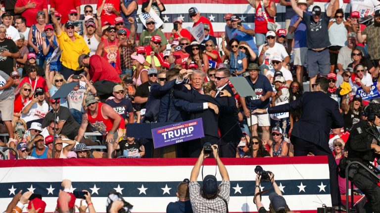 A photograph of Donald Trump surrounded by Secret Service agents on a stage; people in the crowd looking on raise their arms and hold up their cameras.