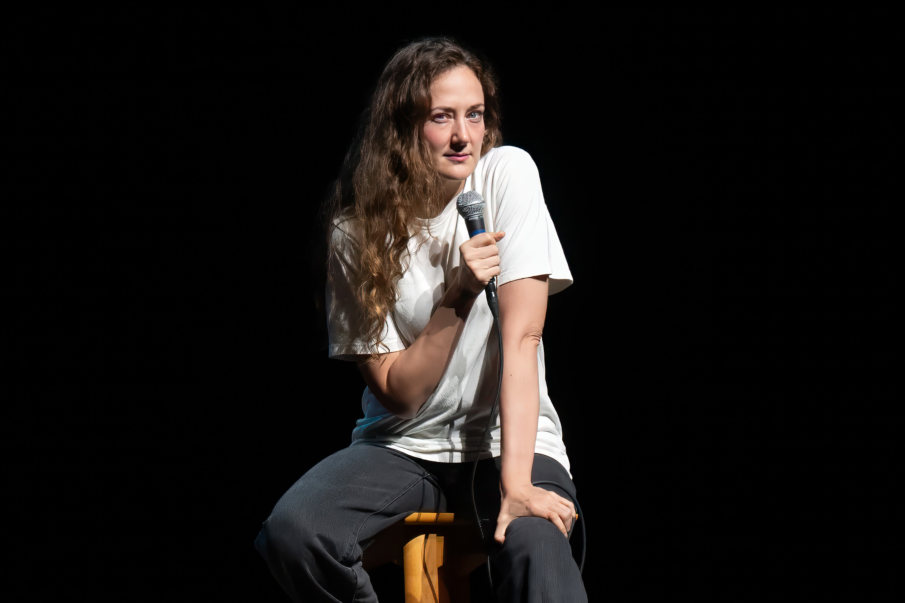 Comedian Jacqueline Novak sits on a stool holding a microphone