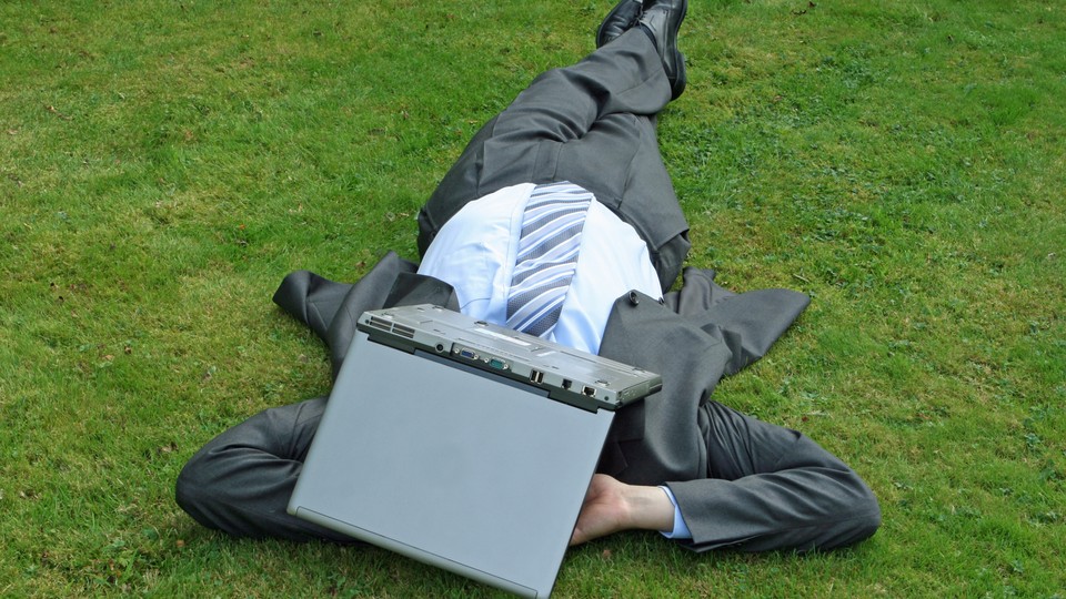 A businessman wearing a suit lays on the grass with a laptop over his face