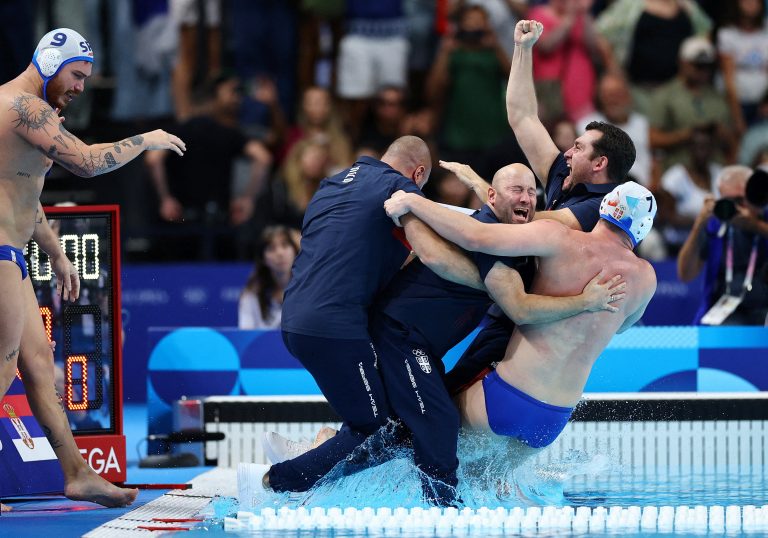 Coaches and a member of a water polo team celebrate, embracing and falling into a pool together.