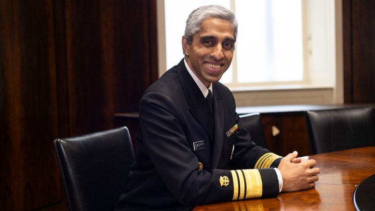 Surgeon General Vivek Murthy, in uniform, grins widely and clasps his hands on a shiny round table