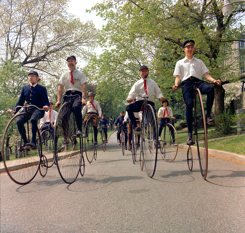 photo of group of men riding old-fashioned bicycles with huge front wheels in a street