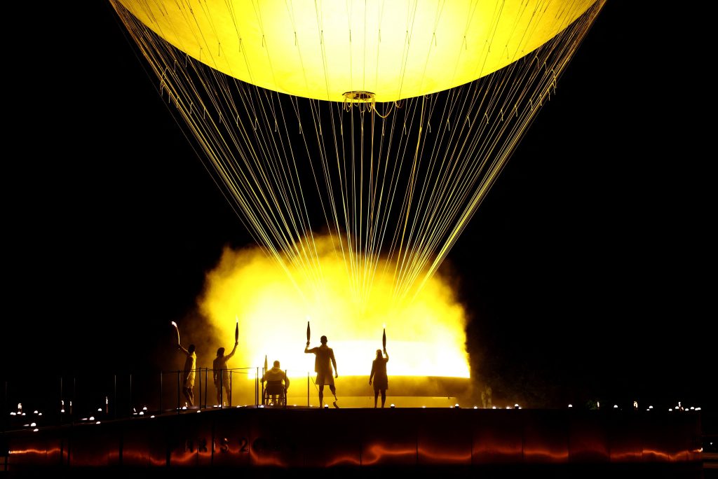 Five torch bearers stand beside a large gondola-like cauldron, below a large balloon, silhouetted by illuminated smoke.