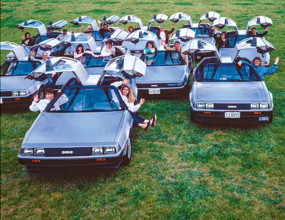 photo of more than a dozen DeLorean cars parked on a grass field, each with both gull-wing doors open and people leaning out and waving