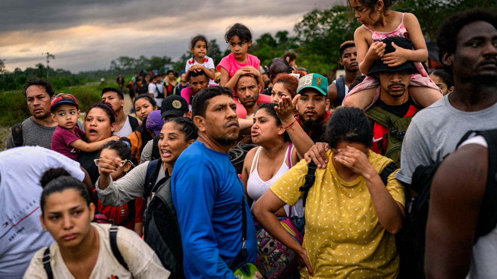 Foto de una fila abarrotada de personas con mochilas, varias de ellas con un niño sobre sus hombros, una mujer con la mano en la frente