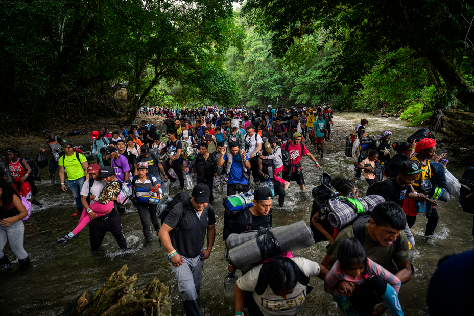 Foto de una multitud de personas caminando por un arroyo rocoso, muchas de ellas llevando niños y equipos, con más multitudes atrás de ellas, todos rodeados por una densa selva.