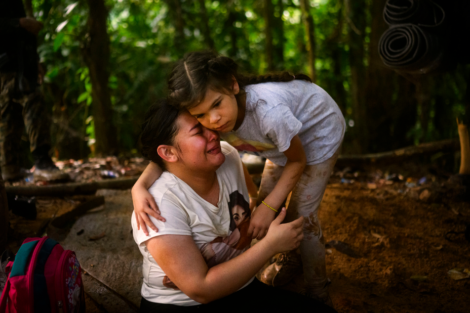 Foto de una niña pequeña parada e inclinada para abrazar a una mujer que llora, sentada en el suelo.