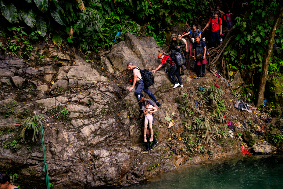 Foto de un hombre que lleva a una niña con las piernas colgando a lo largo de las grietas de una pared de roca muy empinada que cae al agua, con una fila de personas detrás de ellos esperando para intentar también cruzar agarrándose a una sola cuerda horizontal