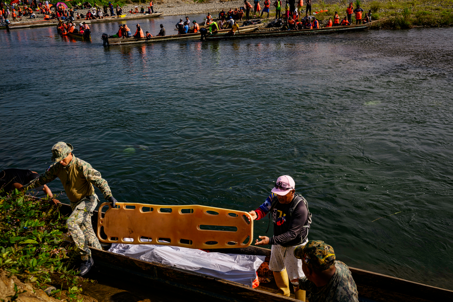 Foto de dos hombres sosteniendo una camilla al lado de un bote muy largo y delgado con una bolsa blanca para cadáveres adentro, con un río y muchos otros botes largos llenos de gente en el fondo