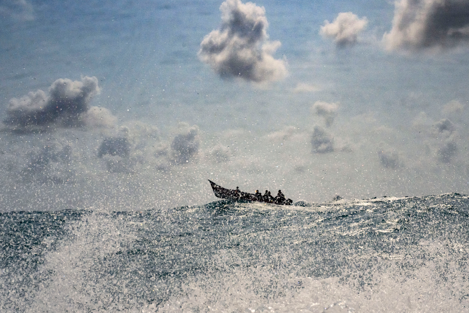 Foto de grandes olas del océano con rocío, con la proa de un bote abierto lleno de gente sobre la cresta de una ola en la distancia