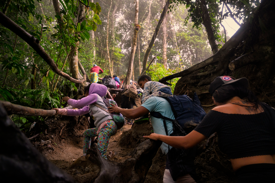 photo of man carrying toddler on shoulders helping small girl up a steep dirt trail holding on to long roots, followed by woman