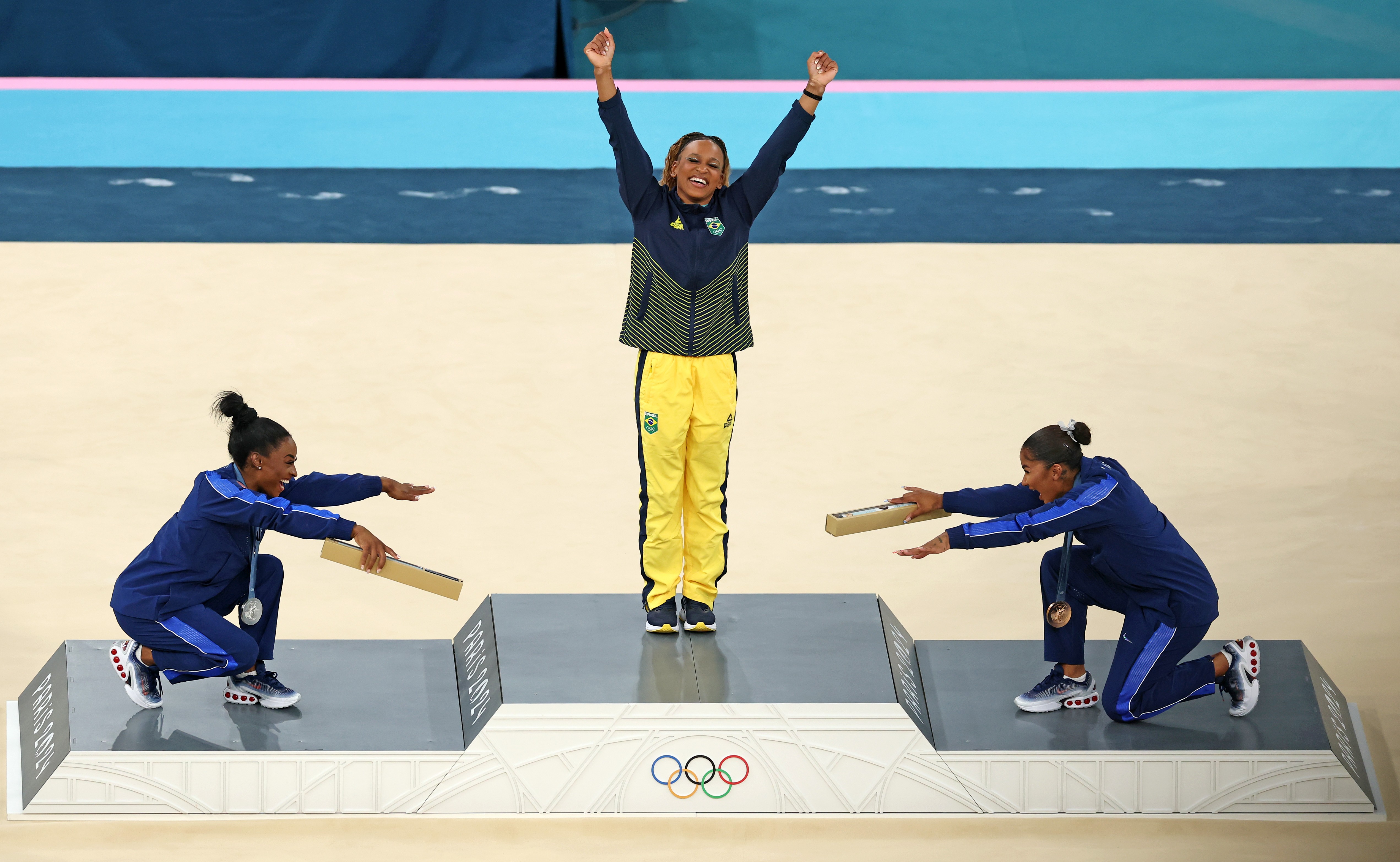 On a medals podium, two gymnasts on either side of the gold medal winner playfully bow down to her, as she raises her arms.