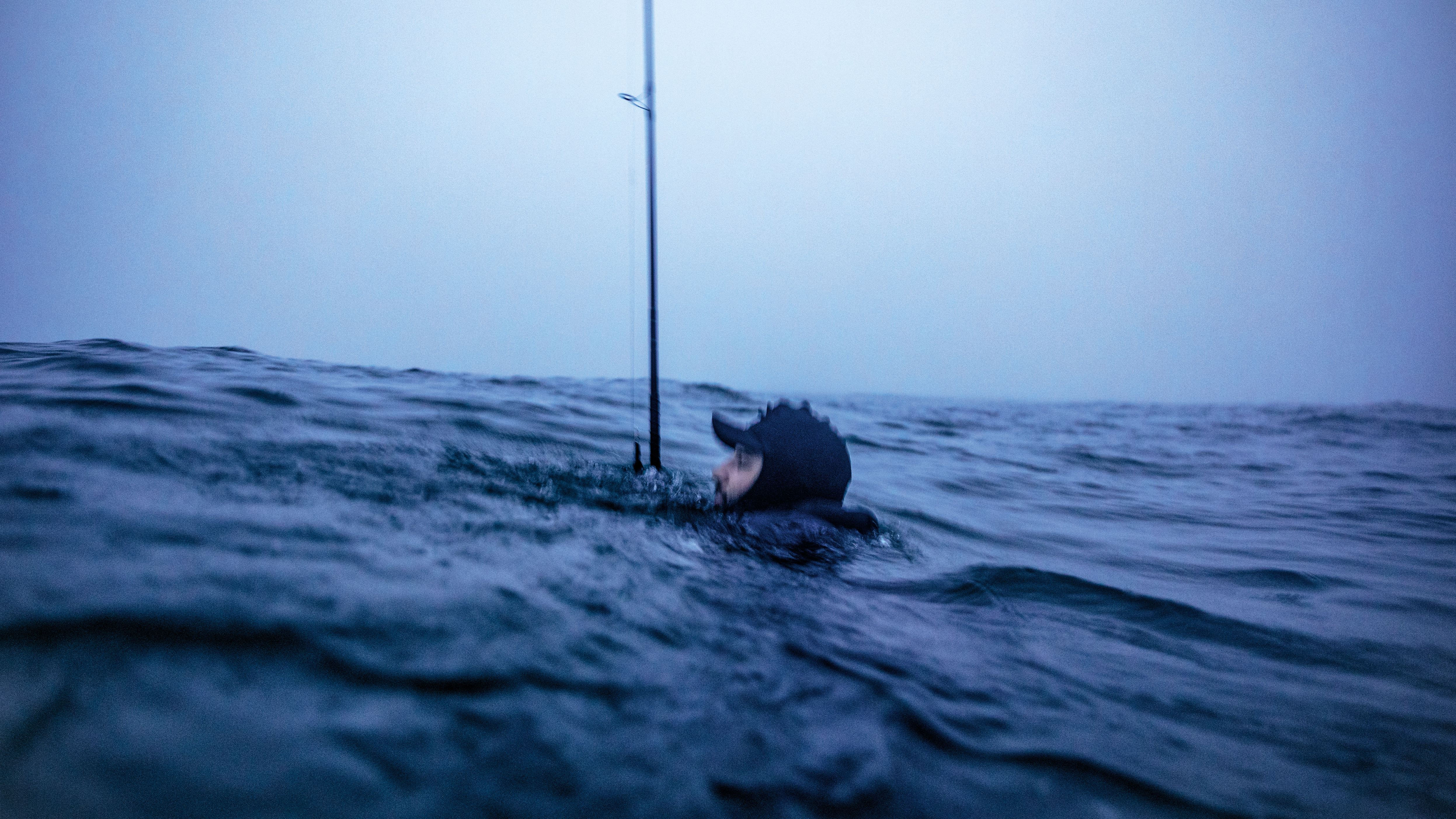 photo of man's head in hat and wetsuit just above an ocean swell, the rest of him submerged, with a large fishing pole vertical in water next to him