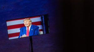 A screen shows Donald Trump speaking in front of an American flag