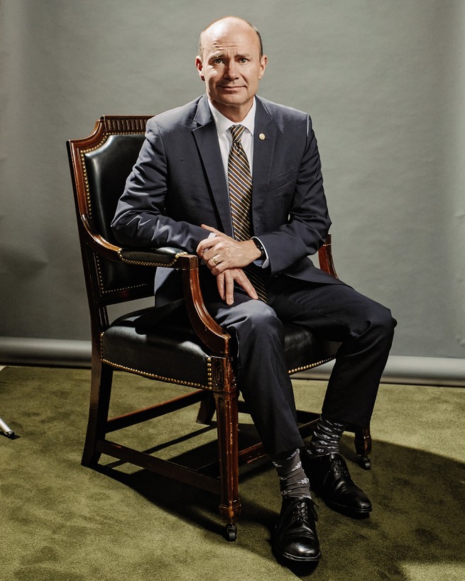 photo of Mike Lee in gray suit sitting in wooden chair on green carpet