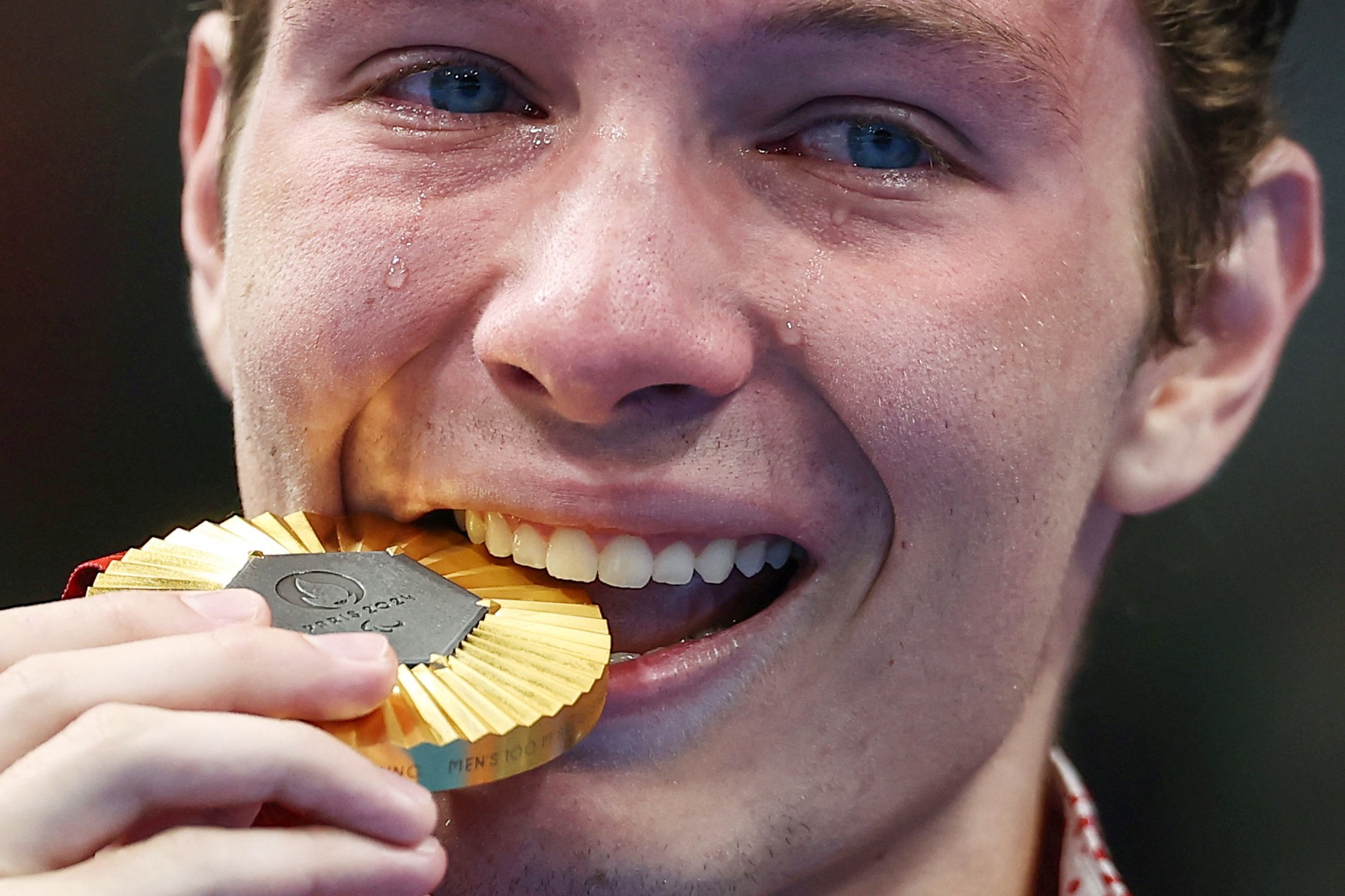 A close view of an athlete's face during a medal ceremony. He is playfully biting a gold medal, with tears falling from both eyes.