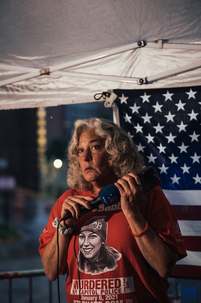 photo of woman wearing Ashli Babbitt t-shirt holding microphone and cell phone under outdoor tent with U.S. flag in background