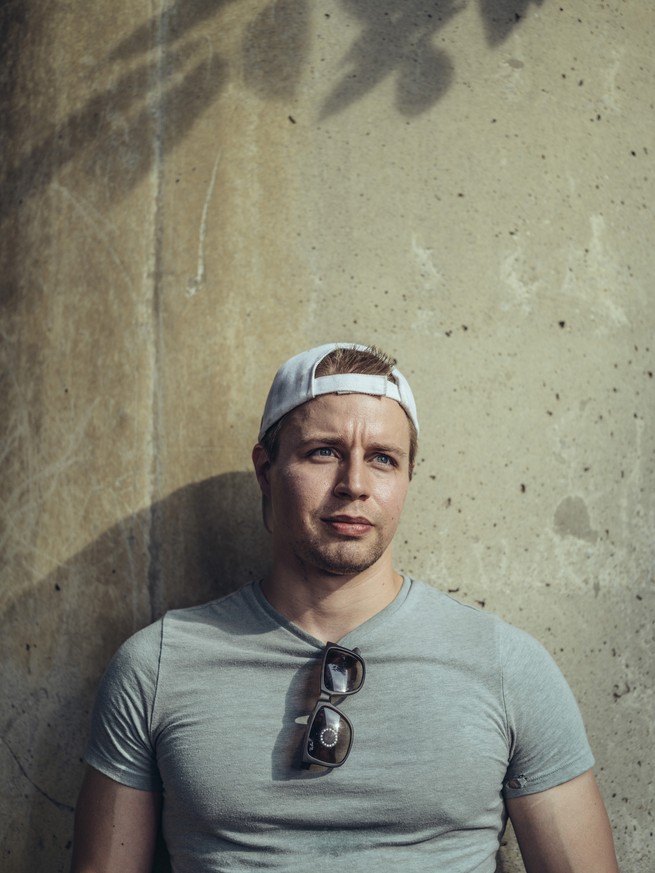 photo of man wearing backwards white baseball cap and gray t-shirt with sunglasses tucked into the collar, standing against concrete wall