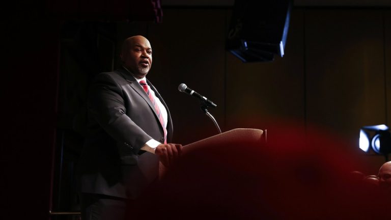 A color photograph of Mark Robinson, in a suit, speaking from a lectern