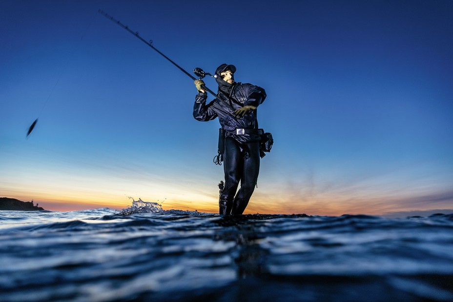 photo from water level at sunrise of man in full-body black wetsuit, jacket, hat, gear belt, boots standing ankle deep on rock in ocean, about to cast with a very large fishing pole baited with fish, with the shore in distant left background