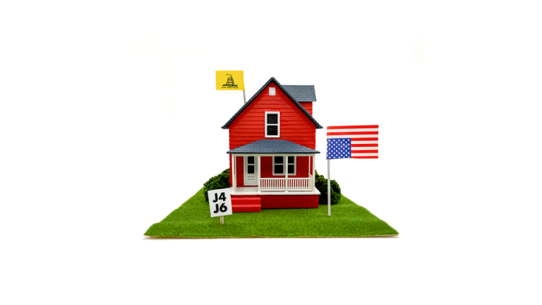 A red toy house on turf with an upside down American flag, J4J6, and a 'Don't Tread on Me' sign on top and in the yard.