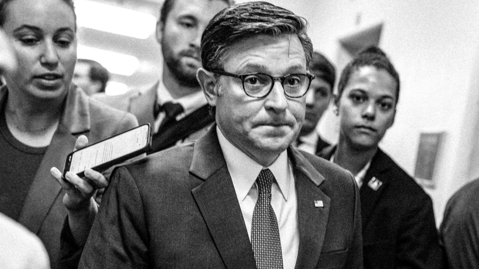 A black-and-white photo of Mike Johnson walking down a hallway, surrounded by other people