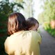 Color photograph of a woman holding a baby, viewed from behind on a wooded path