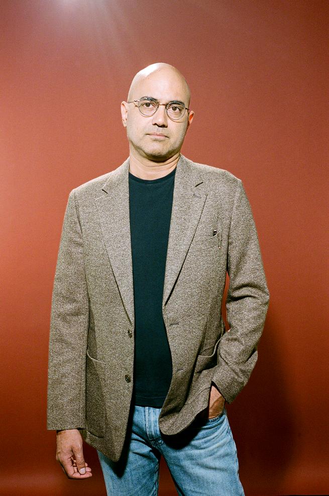 photo of bald man in glasses wearing black t-shirt, tan blazer, and jeans standing with one hand in pocket against brown background