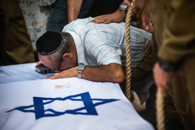 A man in a yarmullke leaning into a casket covered with an Israeli flag (a blue six-pointed star in a field of white).