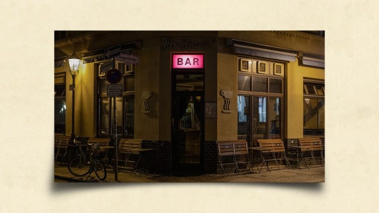 A bar at night, with an illuminated pink sign in the center and folded up tables along its windows