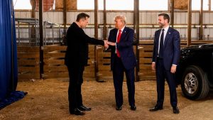 Elon Musk extends his hand to Donald Trump, who pats it. J. D. Vance stands next to them and smiles.