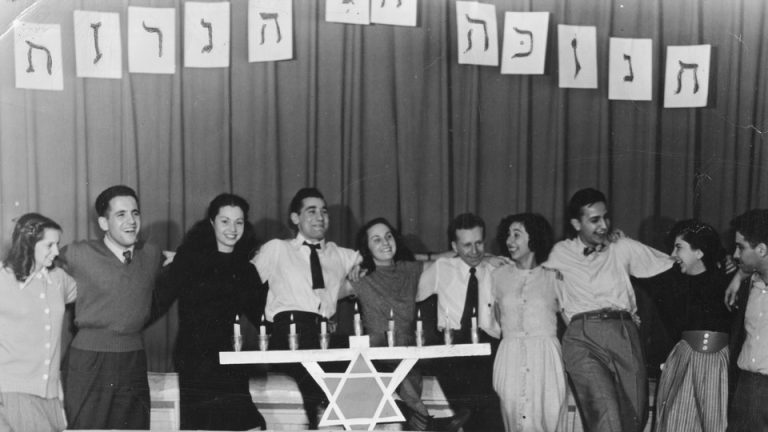 In a black and white photo, a group of men and women laugh and smile in a row standing behind a menorah.