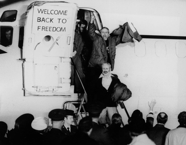 In a black and white photo, American hostages walk out of a plane with arms raised and a sign to their left that says 'welcome back to freedom.'