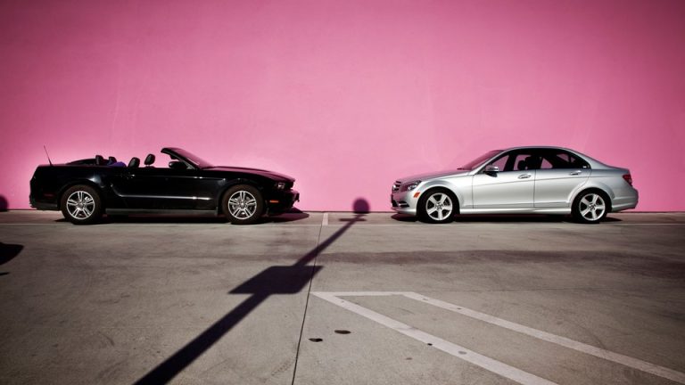 An image of a black convertible (left) and a silver sedan (right) that are parallel parked along a pink wall, facing each other.