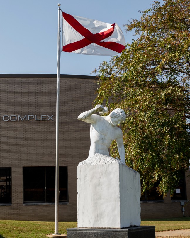 A brown building behind a sculpture and a flag pole of the Alabama flag